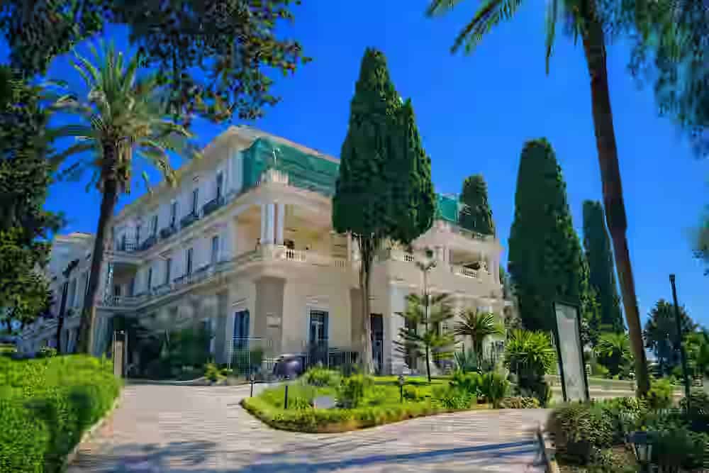 The grand neoclassical facade of Achilleion Palace in Corfu, surrounded by tall cypress trees and palm gardens, easily accessible with a rental car from Corfu Town.