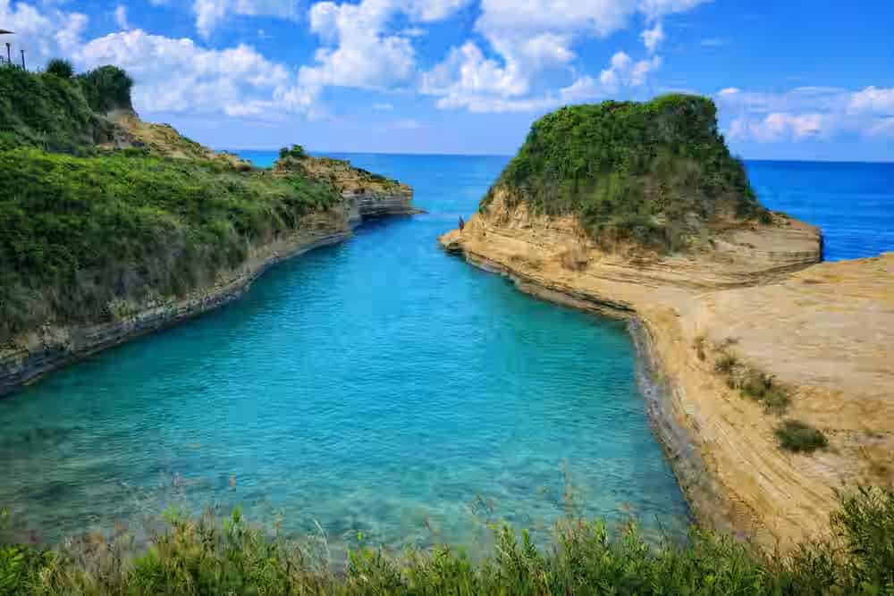 Narrow turquoise sea channel between unique yellow sandstone rock formations and lush green cliffs in Sidari, a top North Corfu attraction to visit with a rental car.