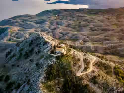 High-angle aerial view from the summit of Mount Pantokrator, showing the monastery, telecommunication antennas, and winding mountain roads overlooking the Ionian Sea coastline.