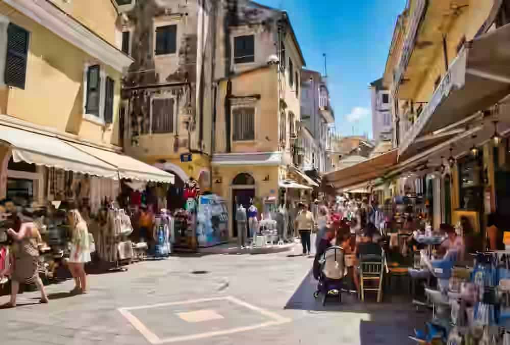 A narrow sunlit street in Corfu Old Town with Venetian-style buildings, local souvenir shops, and outdoor cafe seating, a perfect spot to visit after parking your rental car nearby.