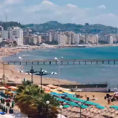 Umbrele colorate pe o plajă cu nisip, cu un ponton lung de lemn și clădirile orașului de coastă Durrës, Albania.