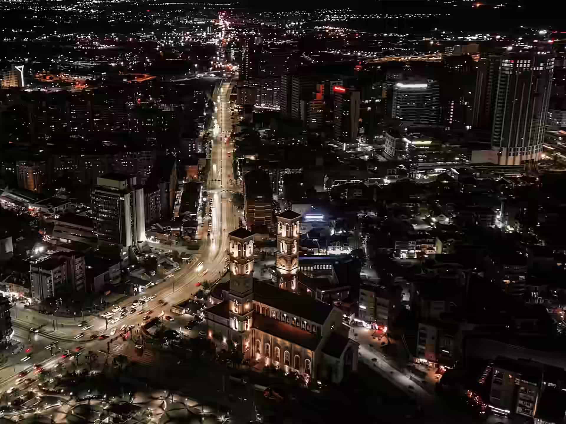 Das Zentrum von Pristina bei Nacht von oben: eine angestrahlte Kirche mit zwei Türmen an einem belebten Boulevard, dahinter beleuchtete Hochhäuser.