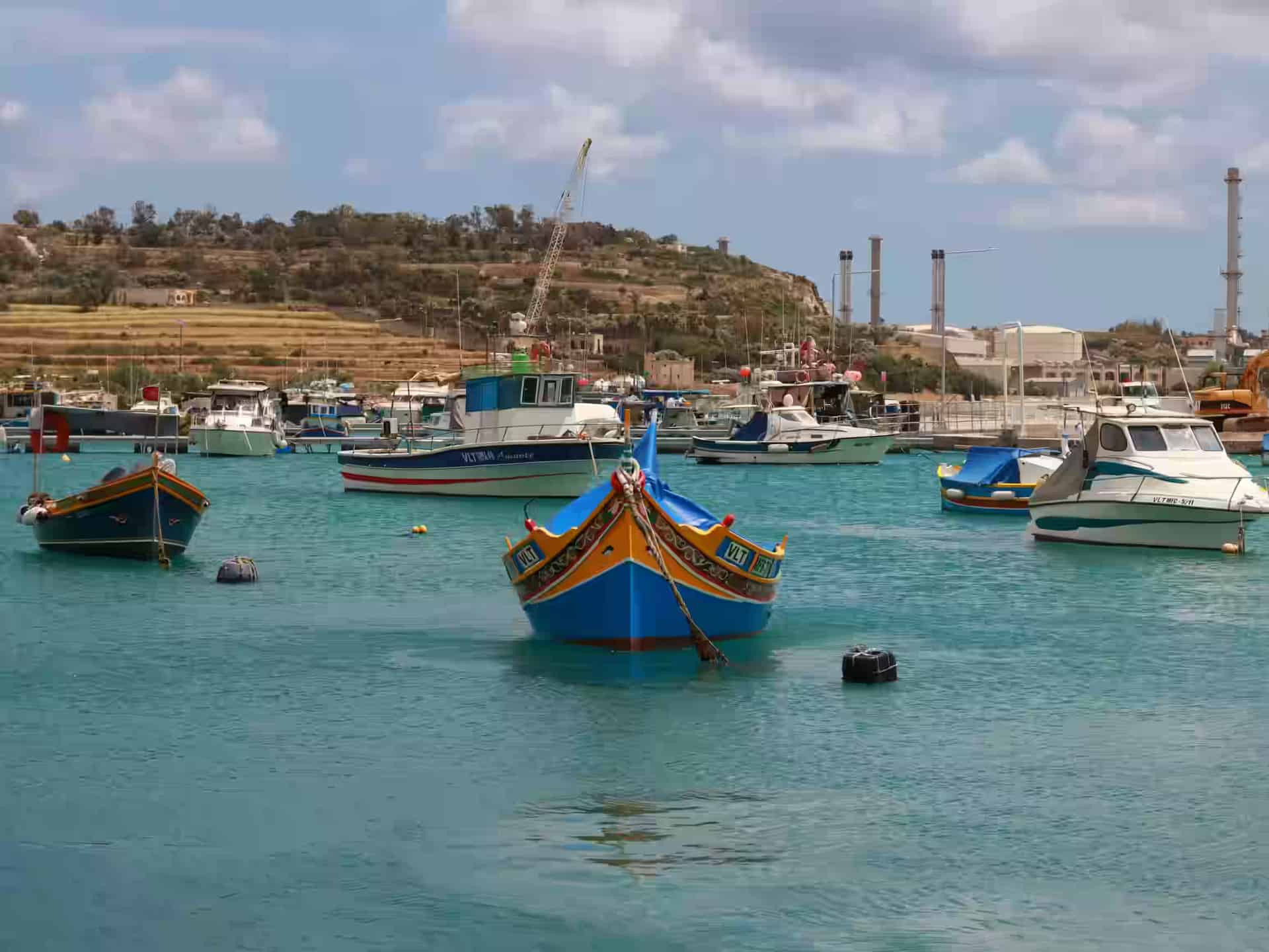 Bunt bemalte Luzzu-Fischerboote im türkisfarbenen Wasser des Hafens von Marsaxlokk, Malta, mit dem Hang über der Bucht dahinter.