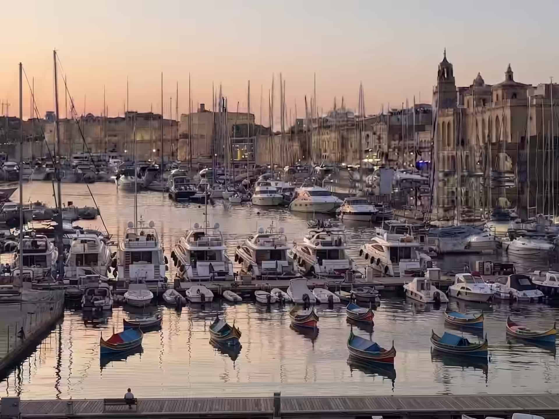 Yachts and painted luzzu boats moored in the Grand Harbour marina at sunset, with the honey-stone waterfront behind