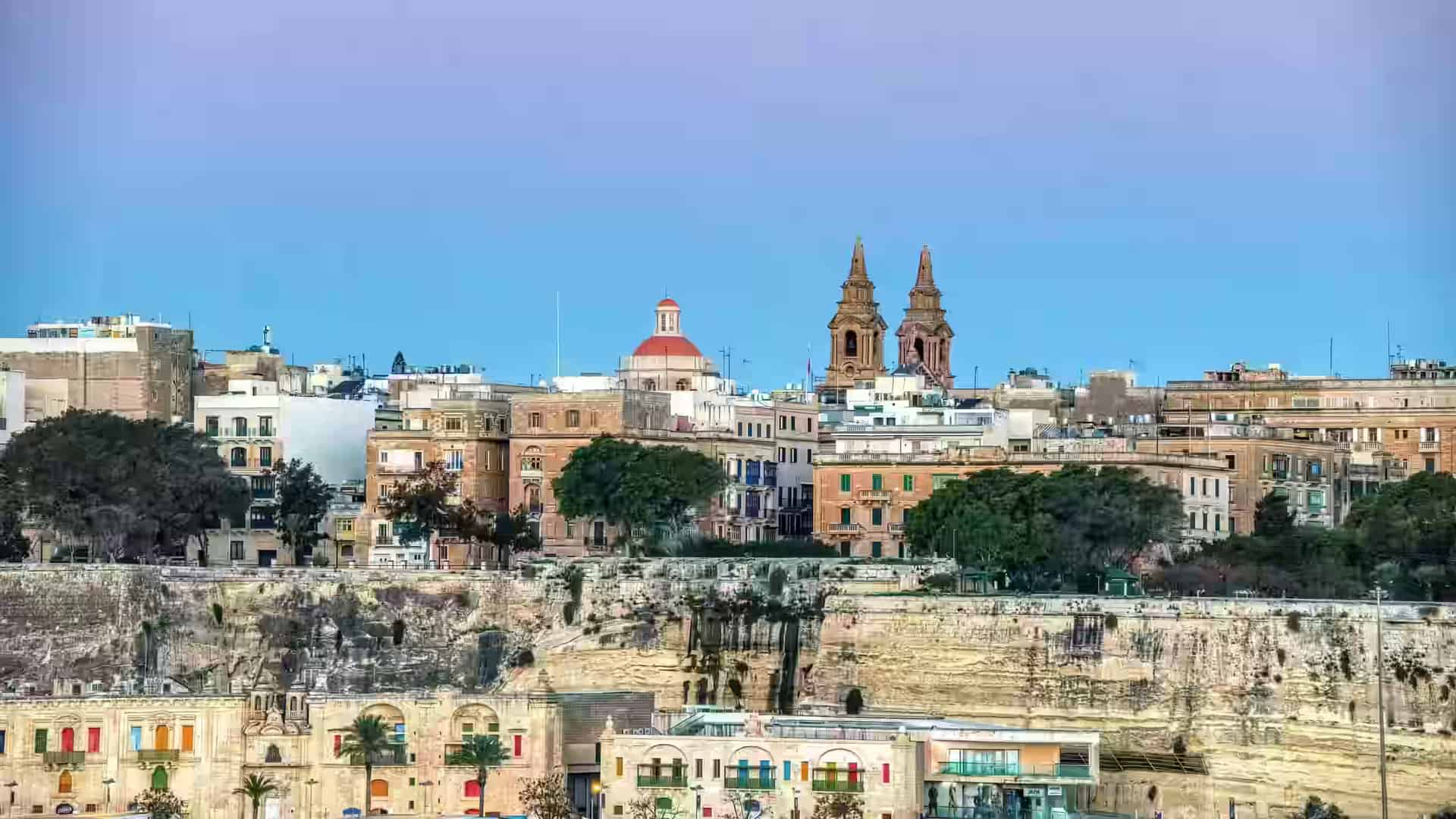 The illuminated skyline of Valletta rising above the stone bastion walls at twilight, with the dome and twin bell towers of the old city against a blue sky