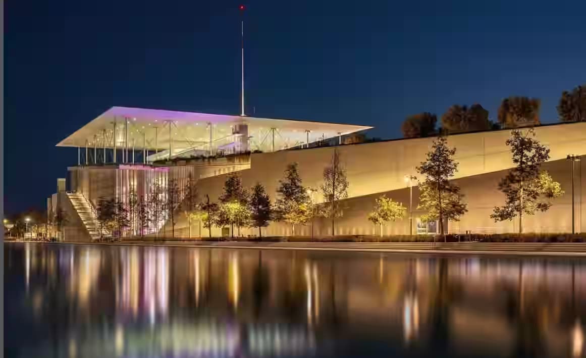 Nowoczesny oświetlony budynek Centrum Kultury Fundacji Stavros Niarchos nocą odbijający się w wodzie kanału.