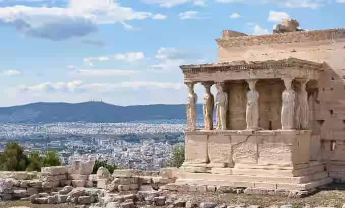 The ancient Erechtheion temple on the Acropolis of Athens featuring the Porch of the Maidens with six Caryatid statues, overlooking the city skyline under a clear blue sky.