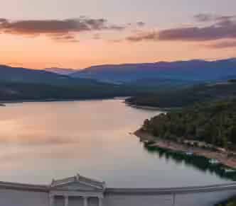Panoramic view of Lake Marathon reservoir and the marble dam at sunset, surrounded by pine trees in Attica, a scenic day-trip destination for car rental travelers from Athens.