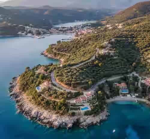 Aerial view of a winding coastal road lined with olive trees and luxury villas leading to Kassiopi harbor, showcasing the scenic northeast Corfu driving route.