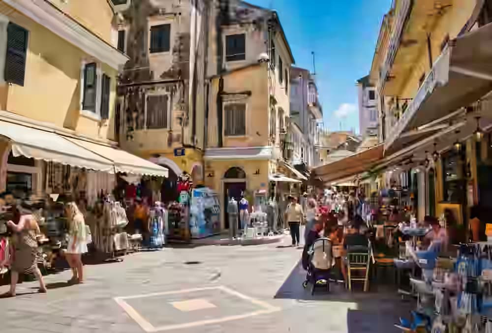Eine sonnenbeschienene Gasse in der Korfu-Altstadt mit venezianischen Gebäuden, Souvenirläden und Café-Terrassen – ideal nach dem Parken des Mietwagens.