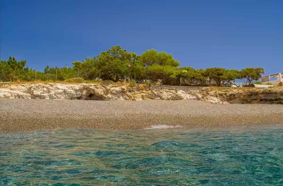 Η παραλία Κουτσουνάρι στην Κρήτη με μικρά πολύχρωμα βότσαλα, τιρκουάζ νερά, βραχώδες πρανές και πράσινες αλμυρίδες κάτω από λαμπερό μπλε ουρανό