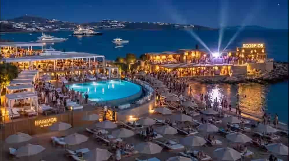 Nächtliche Luftaufnahme eines luxuriösen Beach Clubs auf Mykonos mit beleuchtetem Pool und Yachten im Hintergrund