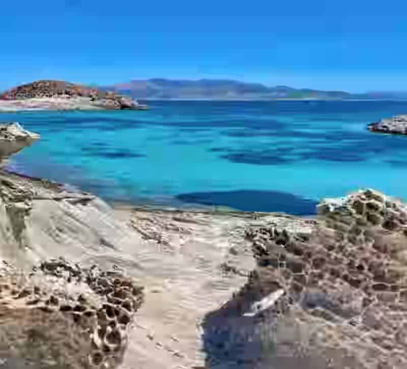 Acque cristalline turchesi e costa rocciosa dell'isola di Antiparo, Grecia, viste dalla strada costiera. Raggiungibile con 7 minuti di traghetto da Pounta, Paros, con il tuo noleggio auto Karpadu.