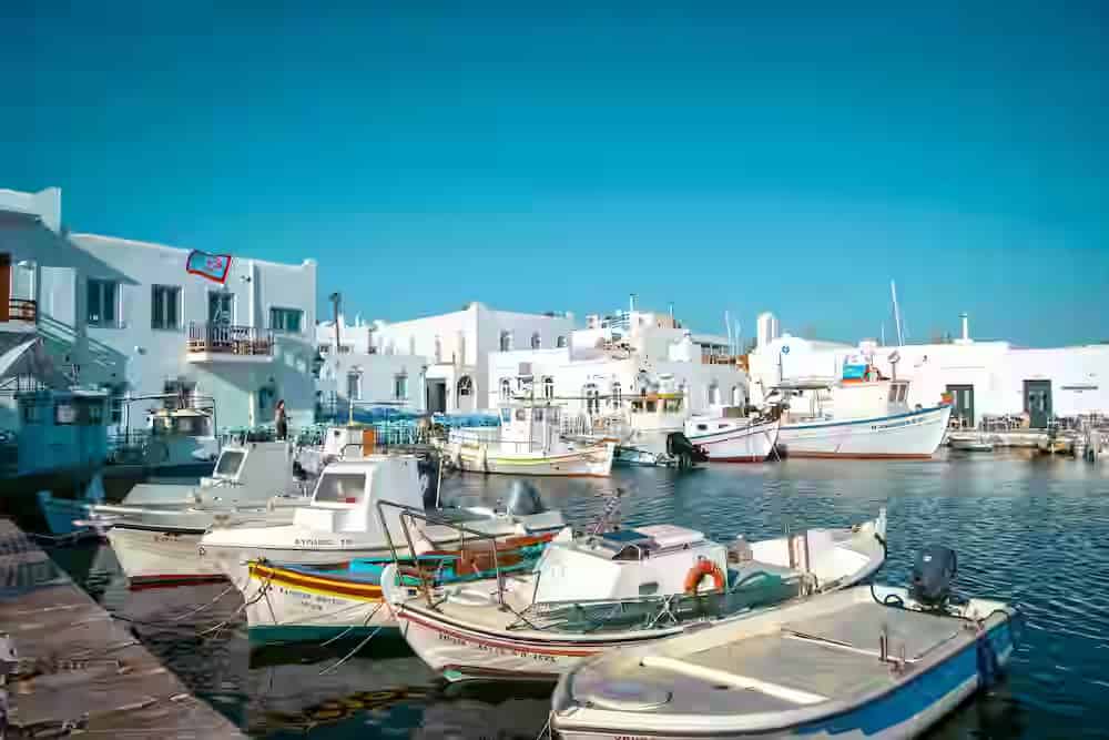 Vista ravvicinata di tradizionali barche da pesca in legno ormeggiate nel vivace porto di Naoussa, Paros, con edifici bianchi sullo sfondo. Scopri il fascino dell'isola con un noleggio auto Karpadu