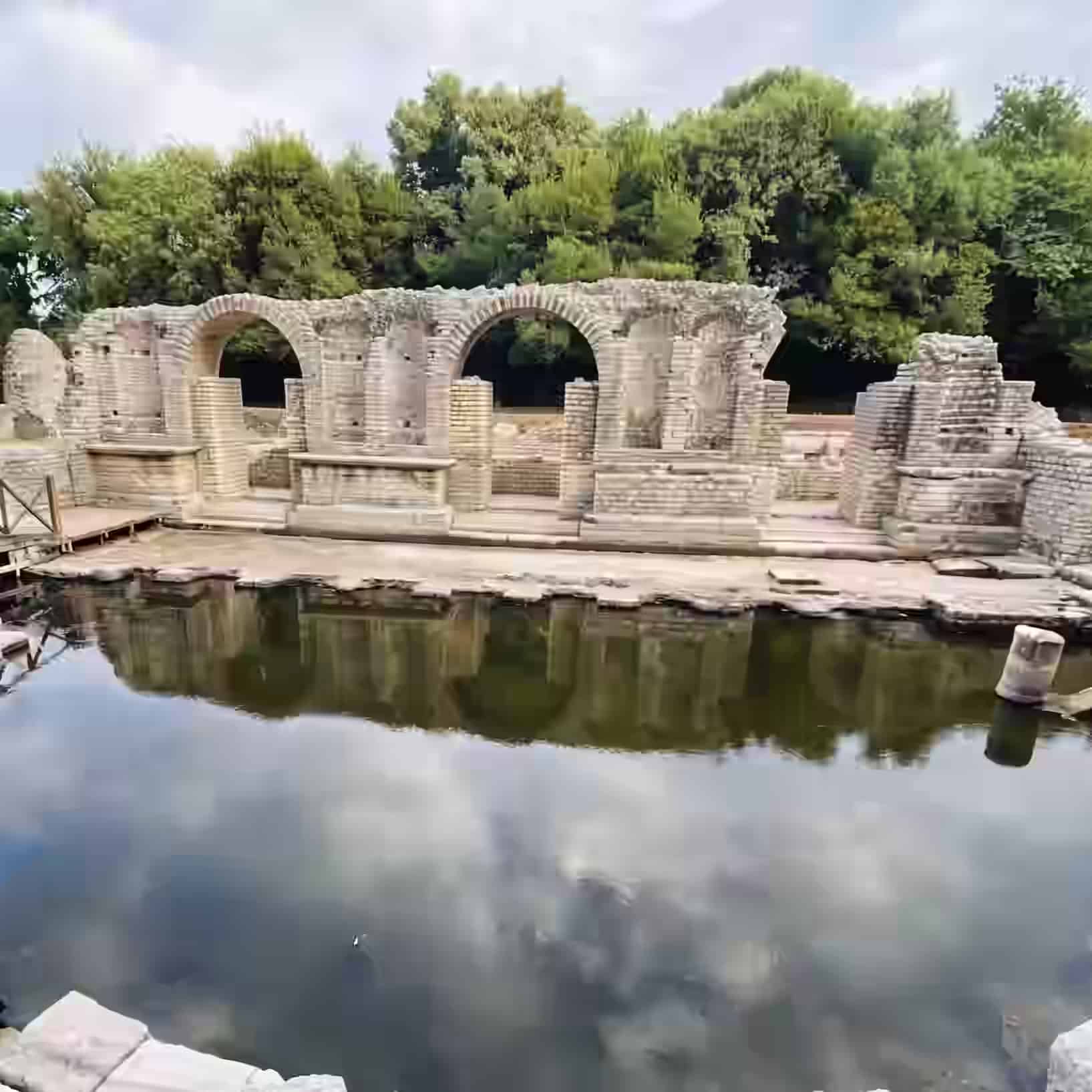 Antike Steinbögen und Ziegelruinen, die sich in einem von Bäumen umgebenen Wasserbecken in Butrint, Albanien, spiegeln.