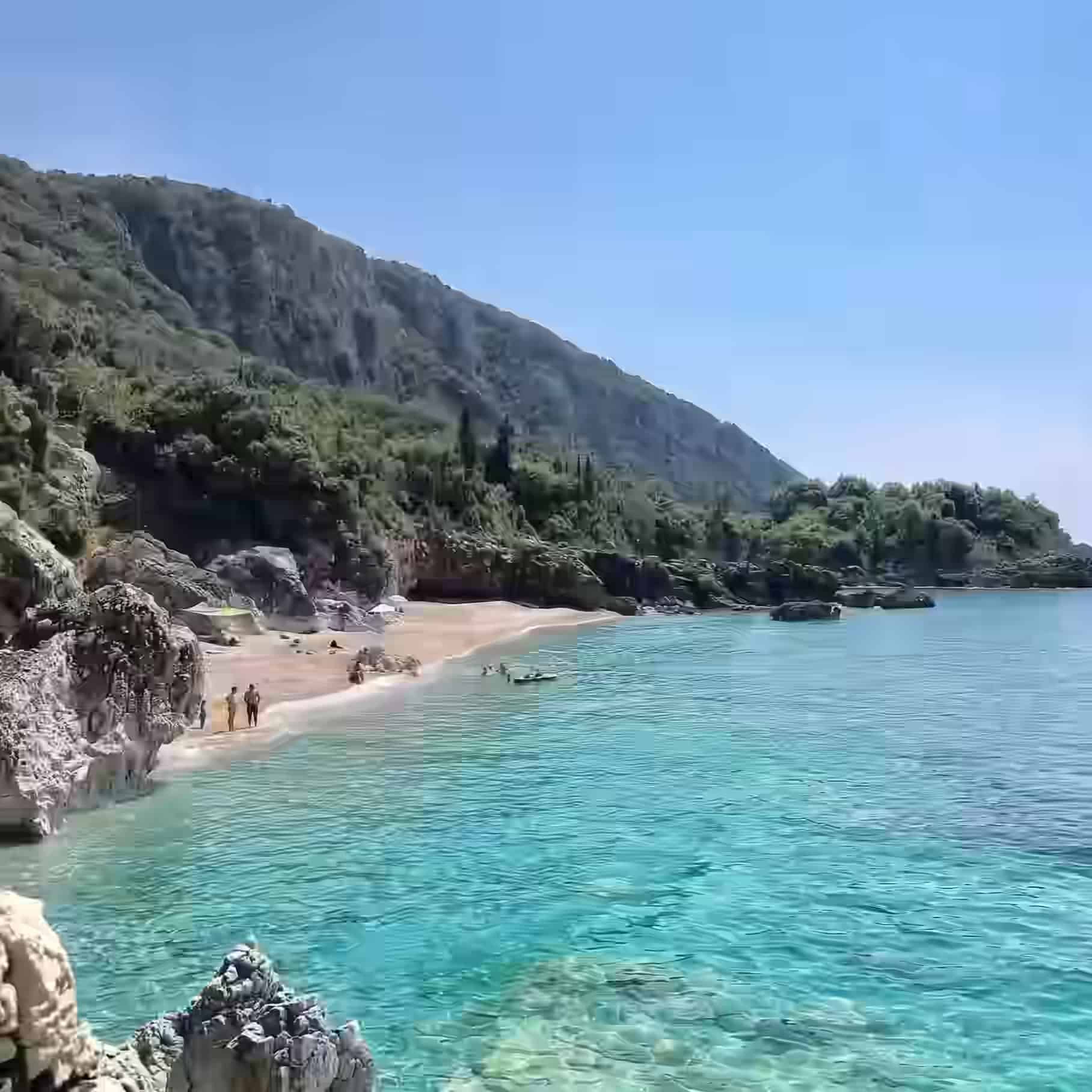 Klares blaues Wasser und ein Sandstrand in einer felsigen Bucht unterhalb steiler grüner Hänge in Dhërmi, Albanien.