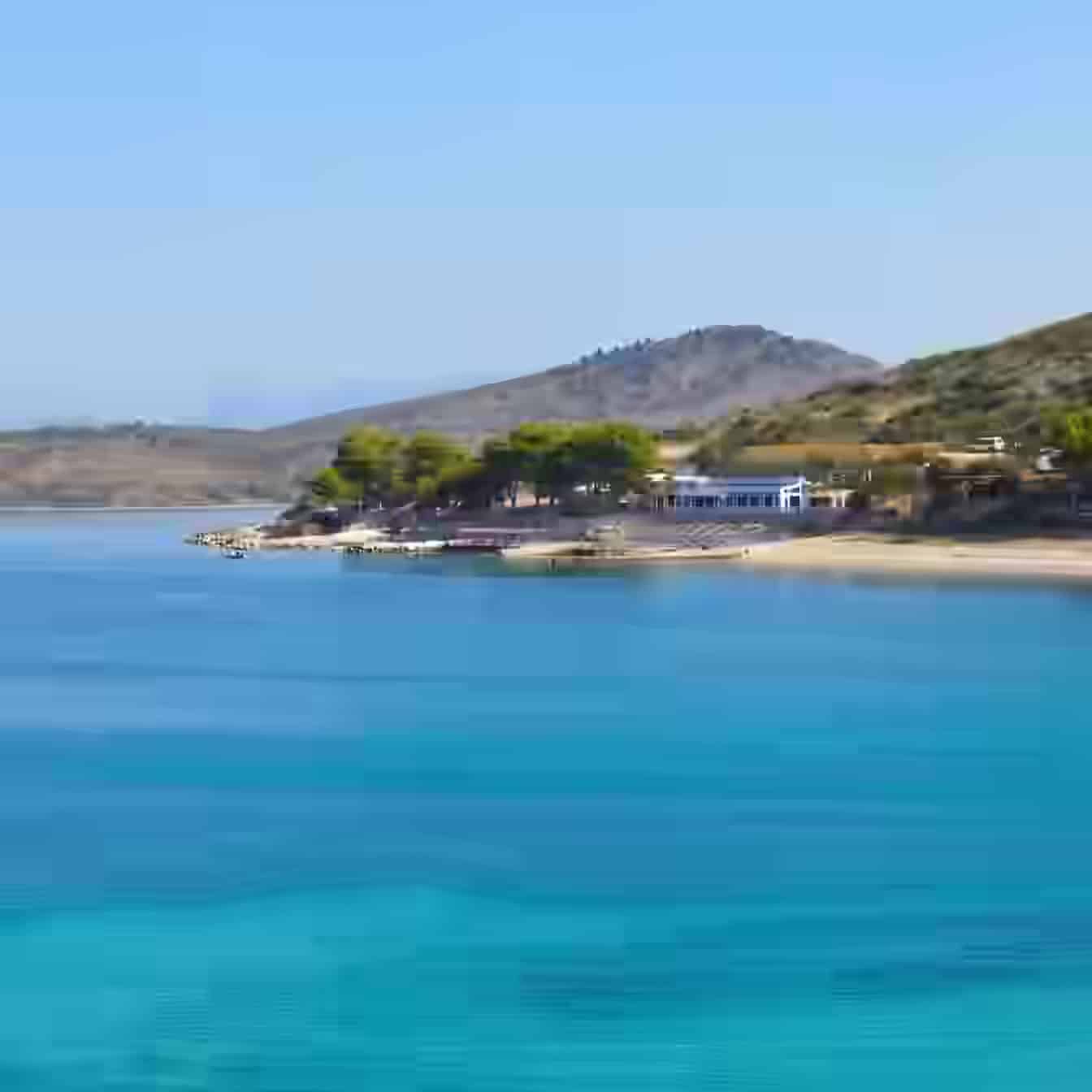 Klares türkisfarbenes Wasser und ein sandiges Ufer, gesäumt von grünen Bäumen, in Ksamil, Albanien, mit sanften Hügeln dahinter.