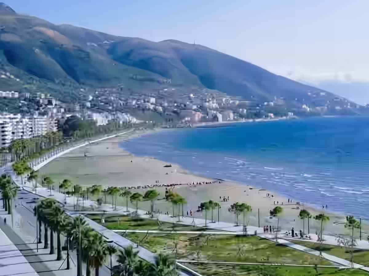 Die von Palmen gesäumte Uferpromenade neben dem breiten Sandstrand und dem blauen Meer in Vlora, Albanien, mit Hügeln im Hintergrund.