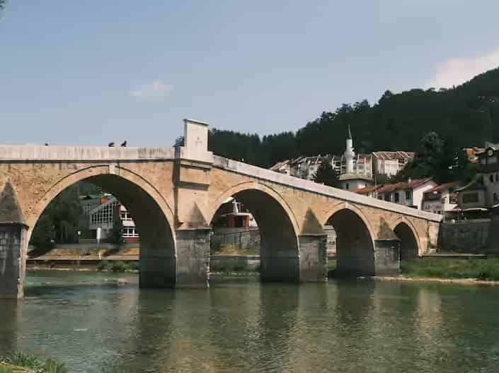 Istorijski kameni most sa više lukova preko zelene reke ispod grada Konjica na padini, Bosna i Hercegovina.