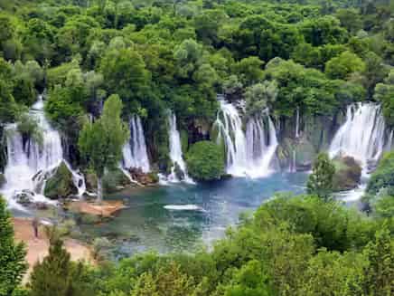 Široki mlazevi vode slivaju se preko zakrivljene krečnjačke litice u zeleno jezerce na Kravici, Bosna i Hercegovina.