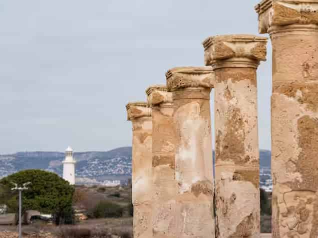 Eine Reihe antiker Steinsäulen mit einem weißen Leuchtturm im Hintergrund im Archäologischen Park von Paphos, Zypern.