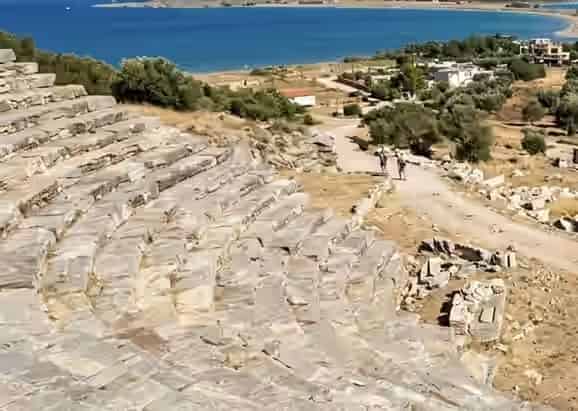 Sièges en pierre des ruines de l'ancien théâtre du site archéologique de Gitana en Grèce, avec un chemin de terre et de l'eau bleue en arrière-plan.