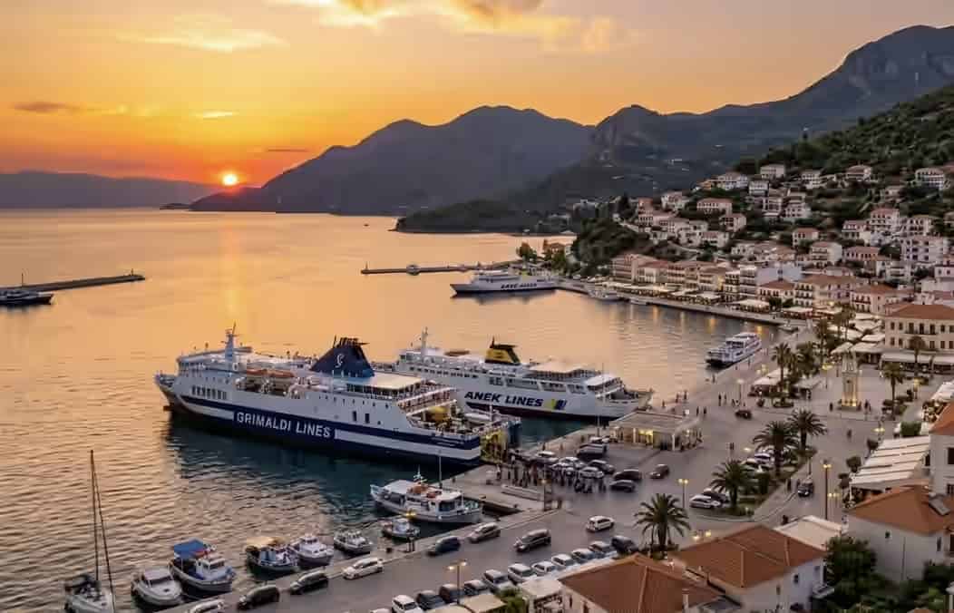 Vue du port d'Igoumenitsa en Grèce au coucher du soleil, avec de grands ferries passagers amarrés, des bâtiments en bord de mer et des montagnes.