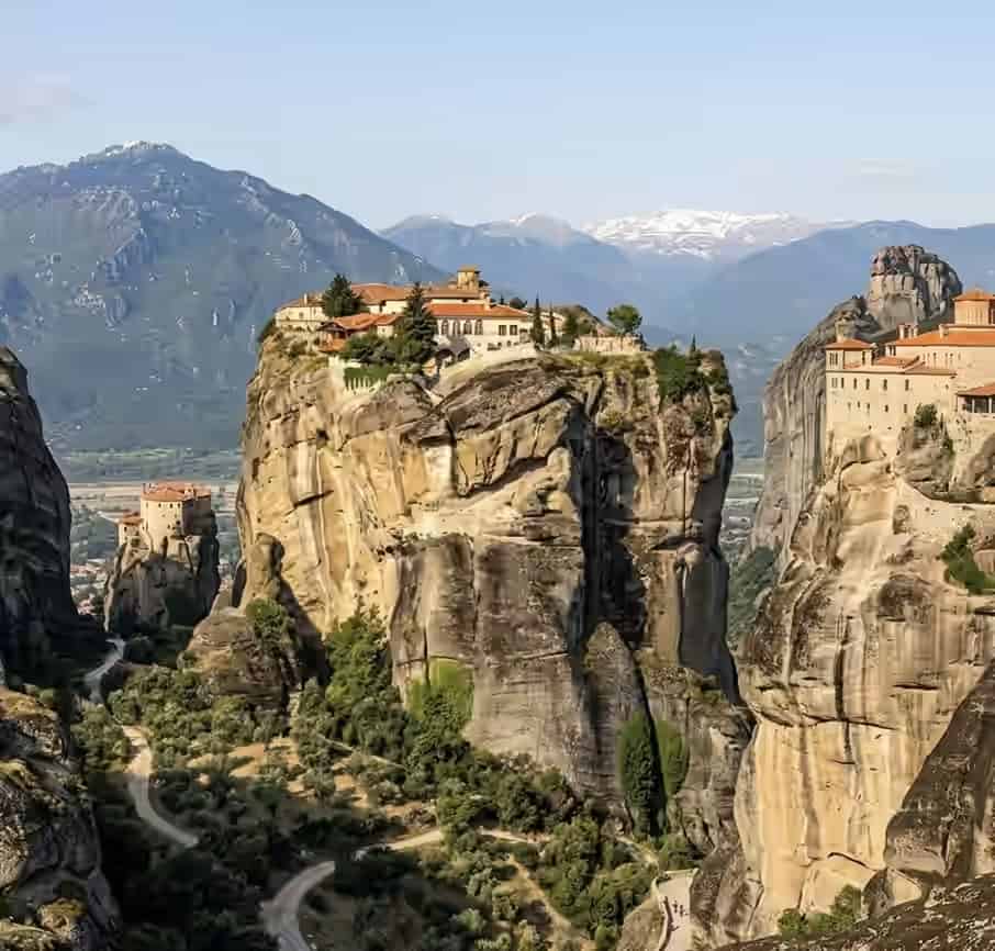 Monastères en pierre construits au sommet d'imposants piliers rocheux à Météores, Grèce, avec une route de vallée sinueuse et des montagnes en arrière-plan.