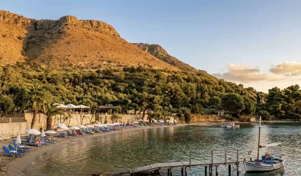 Plage de Plataria en Grèce avec des transats, des parasols blancs, une jetée en bois et un petit bateau amarré devant des montagnes boisées.