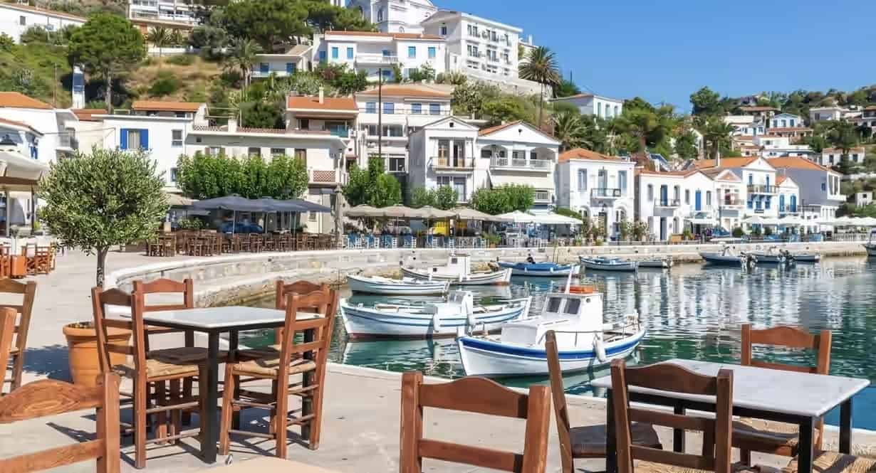 Small fishing boats moored in a village harbor next to outdoor taverna seating in Ikaria, Greece, backed by white hillside buildings.