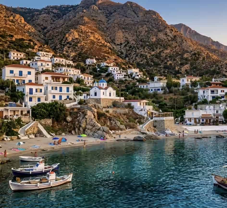 Coastal village of Karkinagri in Ikaria, Greece, featuring white houses on a rocky hillside, a blue-domed church, a small beach, and docked boats.