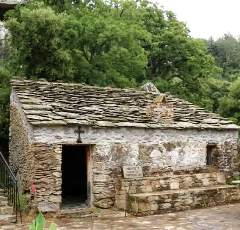 Traditional stone chapel with a slate roof at Theoktistis Monastery in Ikaria, Greece, surrounded by lush green trees.