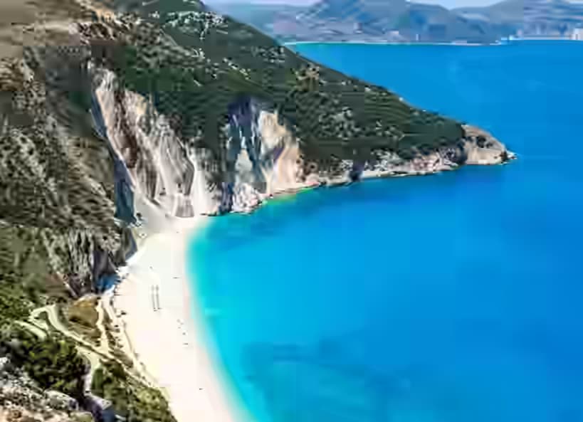 Spiaggia di Myrtos su Cefalonia vista dall'alto con mezzaluna di ciottoli bianchi, falesie calcaree a picco e Mar Ionio blu elettrico