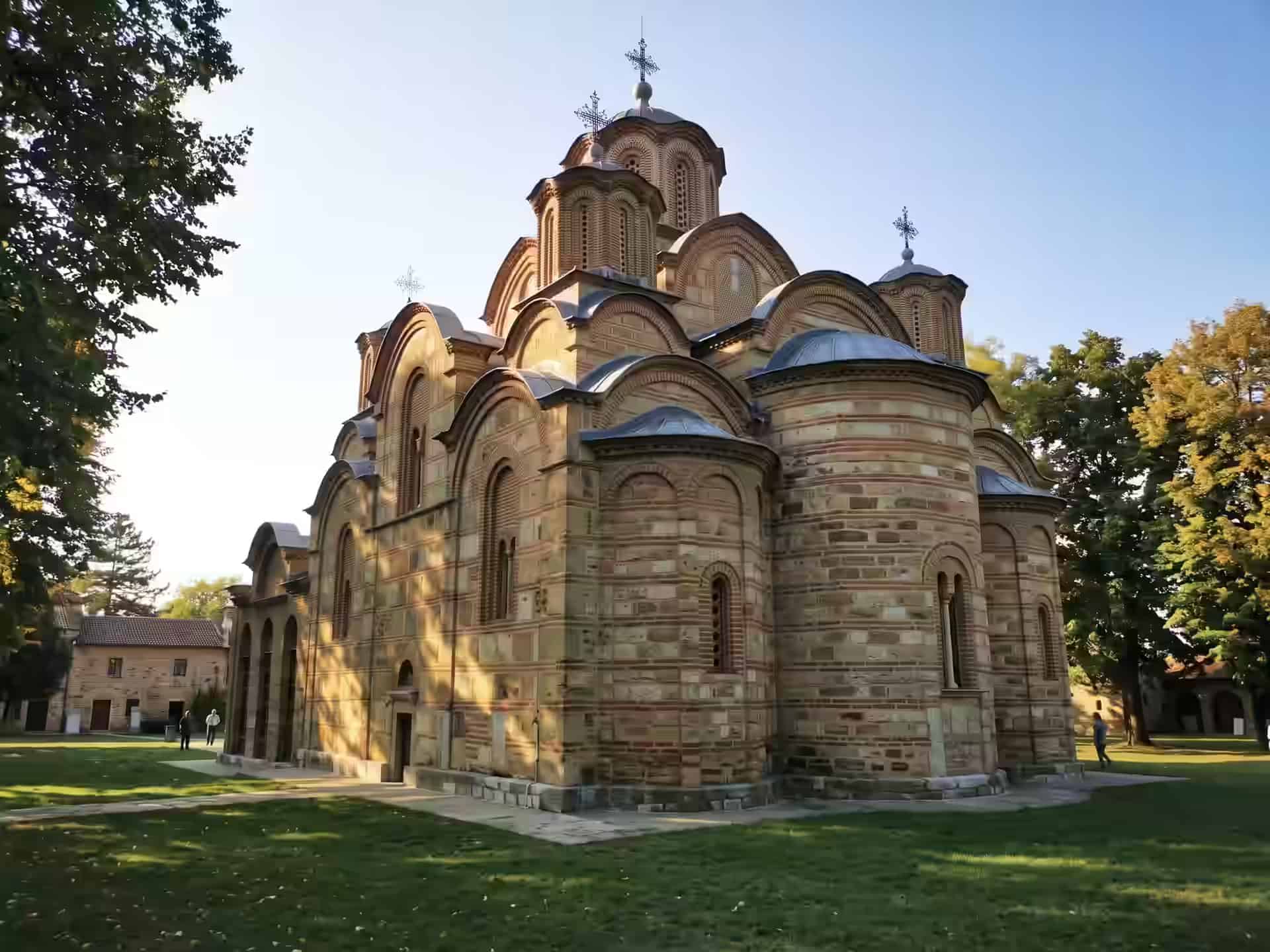 Pięciokopułowa cerkiew klasztoru Gračanica, zbudowana z naprzemiennych pasów kamienia i cegły, na trawniku w popołudniowym słońcu.
