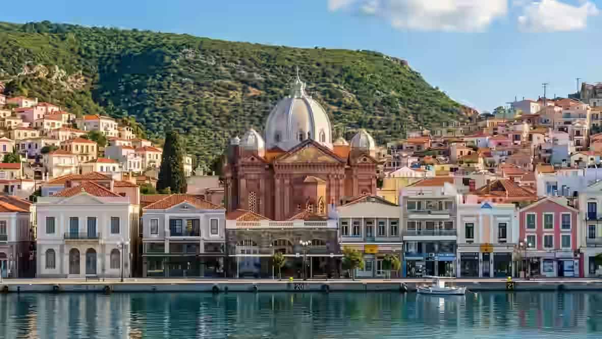 Vue panoramique sur le front de mer de la ville de Mytilène, avec une majestueuse église à coupole s'élevant au-dessus d'une rangée de bâtiments néoclassiques colorés le long du port, tandis que les eaux bleues et calmes reflètent l'architecture vibrante devant une colline verdoyante et luxuriante, à Lesbos, Grèce.