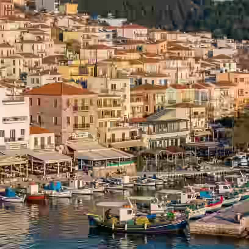 Vue côtière panoramique sur le port de Plomari, avec une jetée en pierre bordée de petits bateaux de pêche en bois, de maisons traditionnelles colorées gravissant la colline et d'une église à coupole imposante devant une toile de fond de collines verdoyantes, à Lesbos, Grèce.
