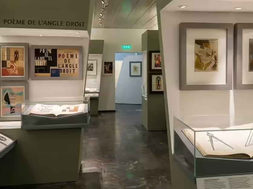 Vue intérieure des expositions modernes du Musée Teriade, présentant des œuvres d'art abstraites encadrées et des vitrines en verre contenant des livres ouverts, devant des cloisons de galerie vert olive, à Varia, Lesbos, Grèce.