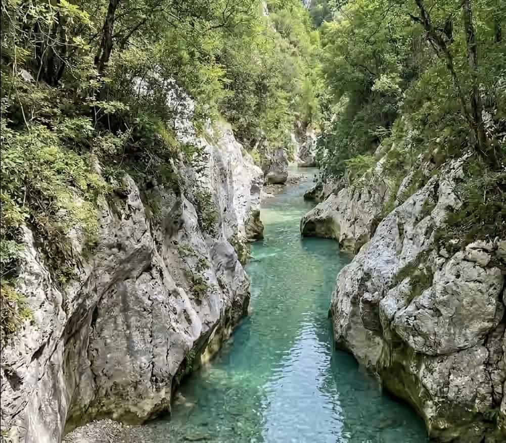 A narrow section of the Glyki Gorge in Greece, with clear turquoise river water flowing between steep white rock cliffs topped with green trees.