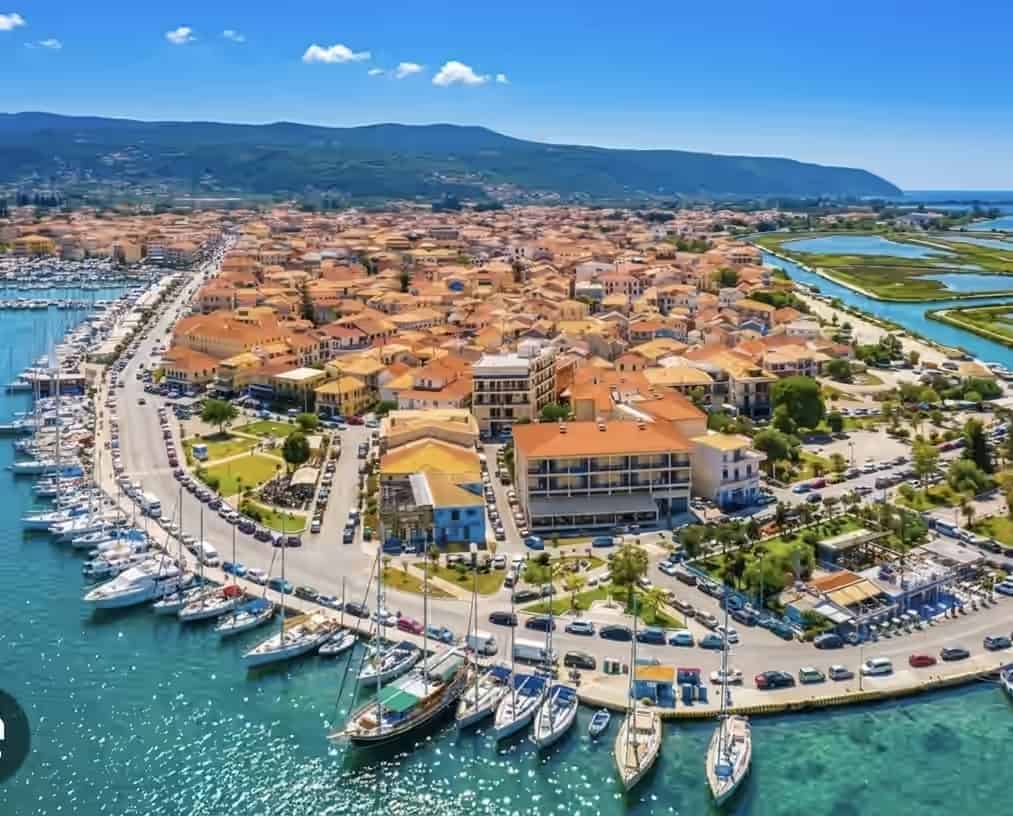 Aerial view of Lefkada town in Greece, showing a curved marina with sailboats, a waterfront road, and buildings with orange tiled roofs.