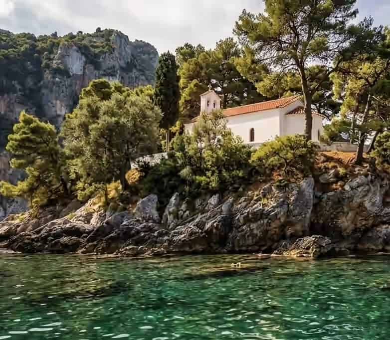 A white chapel with a red-tiled roof and pine trees on a small rocky islet in the clear green waters of Parga, Greece, backed by steep cliffs.