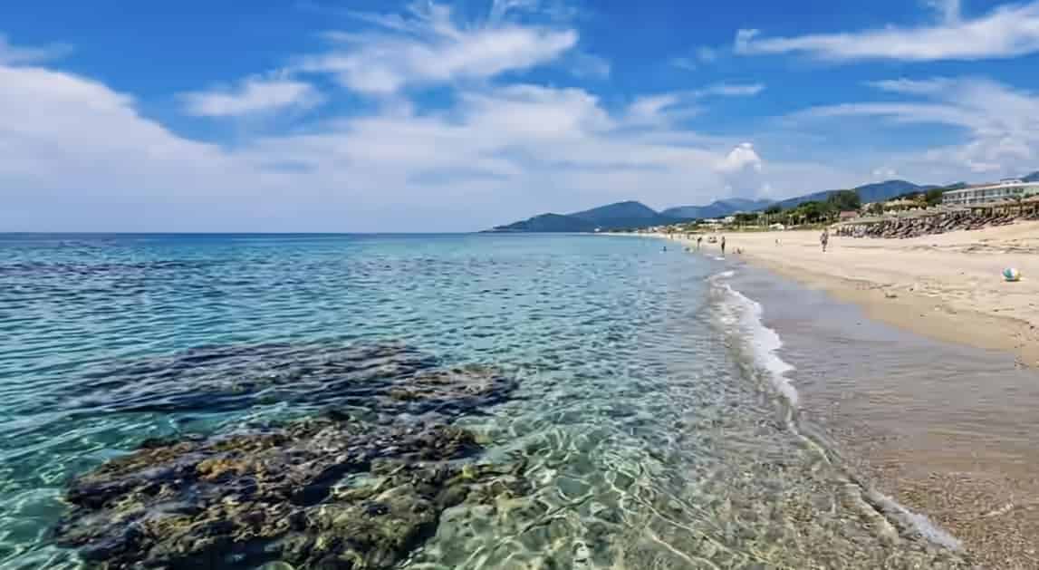 View along the sandy shoreline of Monolithi beach in Preveza, Greece, with clear shallow water, rocks in the foreground, and distant mountains.