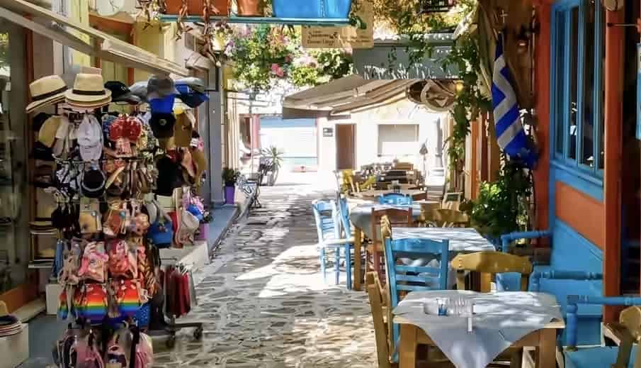 A cobblestone alleyway in the old town of Preveza, Greece, with colorful outdoor souvenir displays and traditional blue taverna chairs under leafy vines.