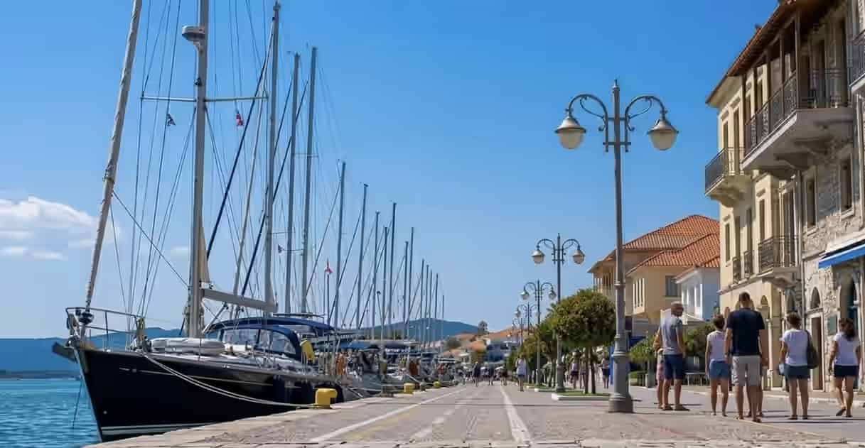 People strolling along the waterfront promenade in Preveza, Greece, with a row of docked sailboats on the left and traditional buildings with balconies.
