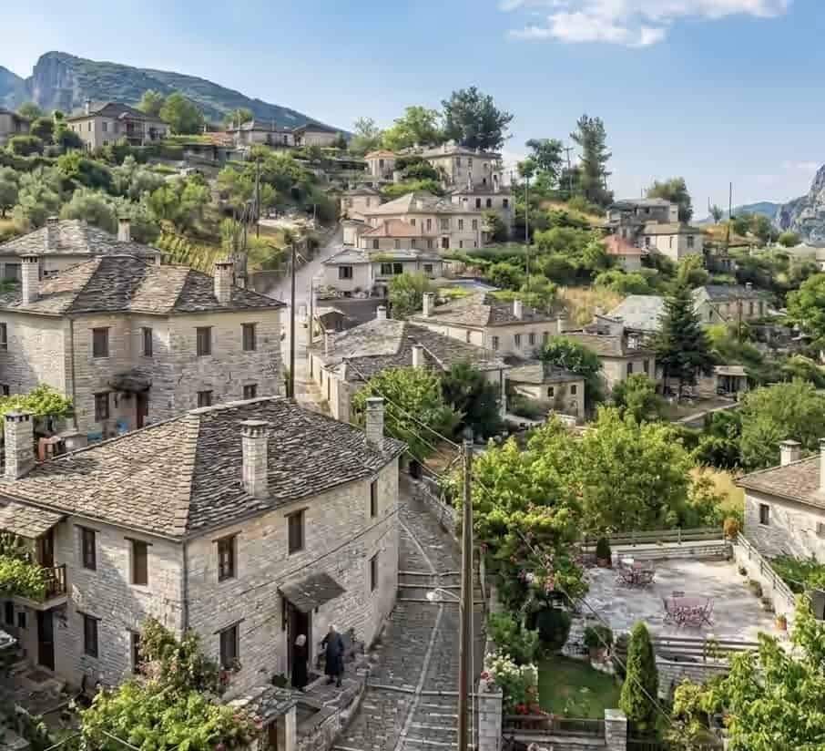 Traditional village in Zagorohoria, Greece, with characteristic stone houses and dark slate roofs along a cobblestone path on a green hillside.
