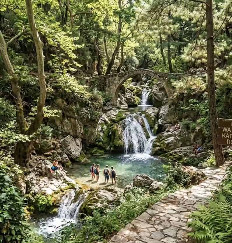 People wading in the natural pools of Potami waterfalls in Samos, Greece, surrounded by a lush green forest and an arched stone bridge.
