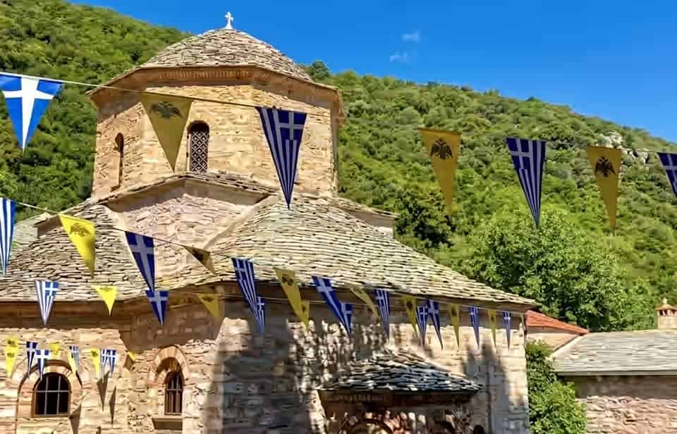 Stone church of Evangelistria Monastery in Skiathos, Greece, decorated with Greek and Byzantine flags against a green forested hillside.