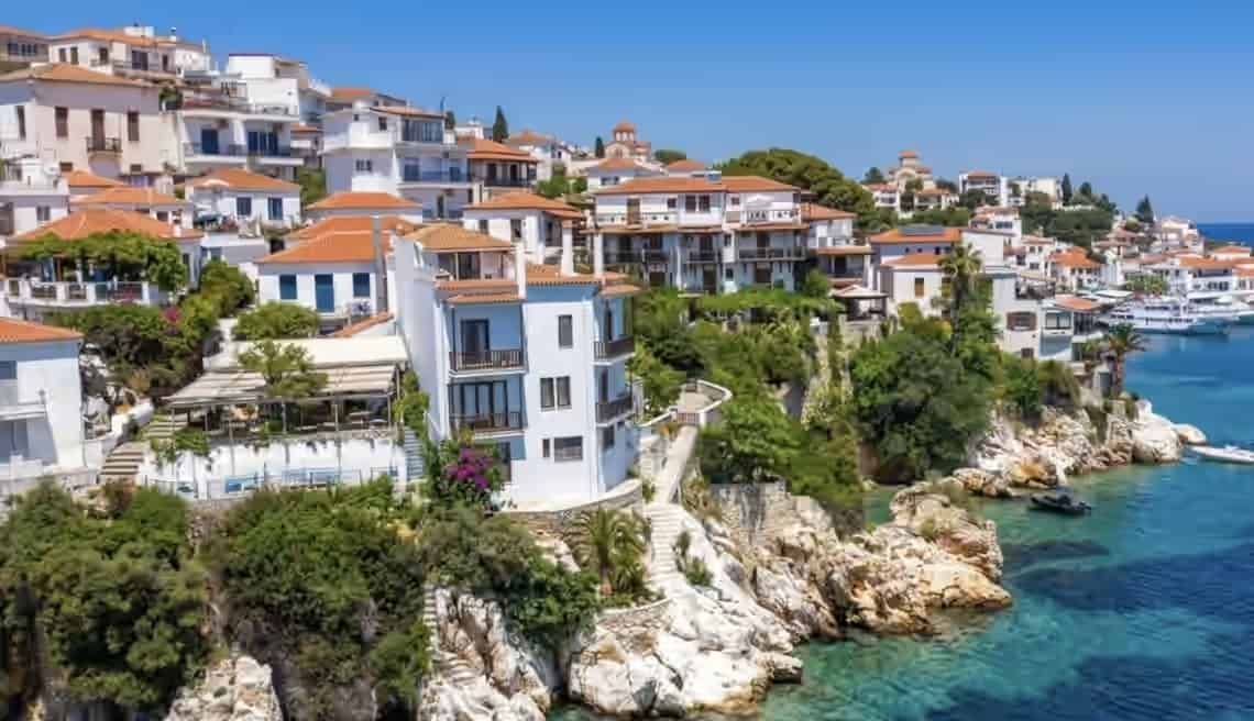 Scenic view of Skiathos Town in Greece, featuring traditional white buildings with red-tiled roofs on a rocky hillside above clear blue water.