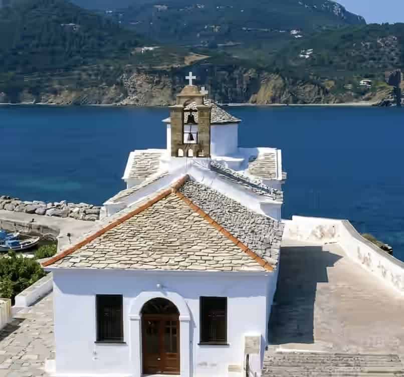 A traditional white church with a stone roof and bell tower overlooking the blue sea in Skopelos, Greece, with rugged coastline in the background.