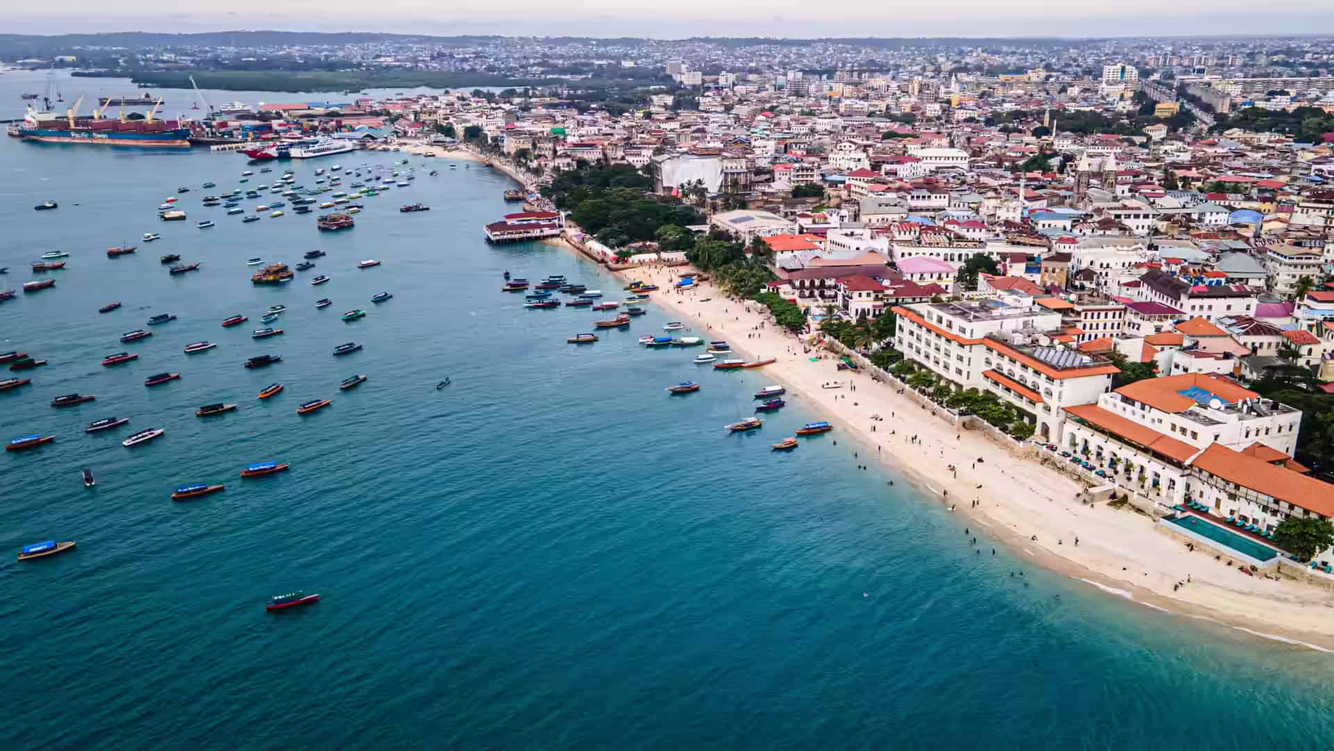 Luftbild der Uferfront von Stone Town: Dutzende Holzboote in der Bucht vor Anker, ein Sandstrand und Uferhäuser mit roten Dächern, dahinter der Hafen.