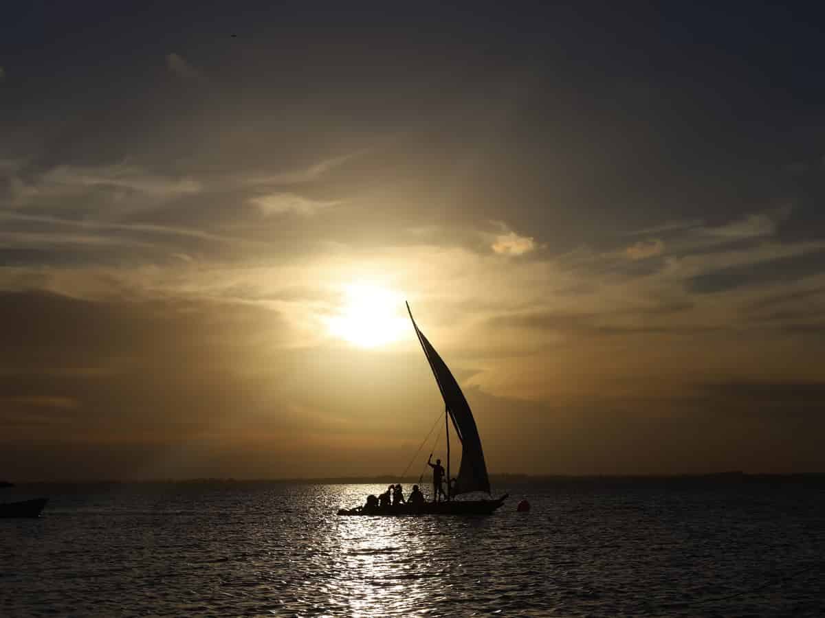 Eine Dhau unter Segeln mit Passagieren an Bord, als Silhouette vor einem goldenen Sonnenuntergang vor Sansibar.